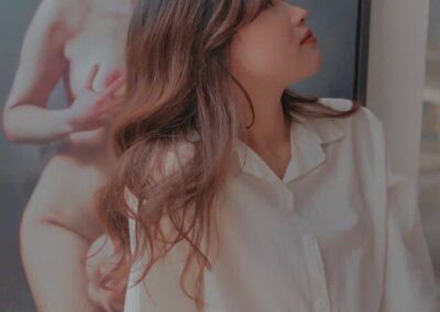 Nude woman with long wavy hair and artistic makeup, posing in front of a large art piece.