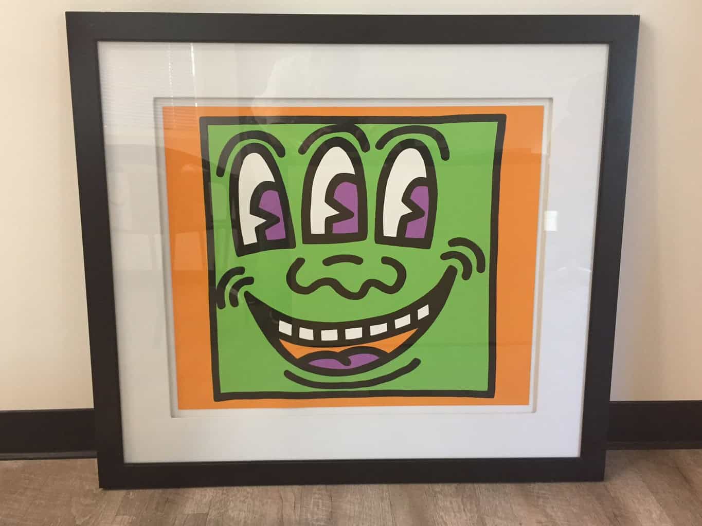 A vibrant illustration of Keith Haring's iconic Three-Eyed Monster symbol, positioned prominently in the foreground, characterized by bold outlines and a playful, cartoonish style. This monster features three expressive eyes and a wide, devious grin, embodying themes of capitalism and desire. In the middle ground, incorporate a stylized flying devil with outstretched wings and mischievous energy, juxtaposed with a serene angel, symbolizing transcendence and balance. The background showcases a swirling blend of bright colors, reflecting Haring&rsquo;s exuberant style, illuminated by a warm, golden light that evokes a sense of hope amidst chaos. Use a slightly tilted perspective to create dynamic tension within the composition. The overall mood is energetic and thought-provoking, capturing Haring's commentary on society. Logo of "Poppins Shop" subtly integrated into the design without dominating the artwork.